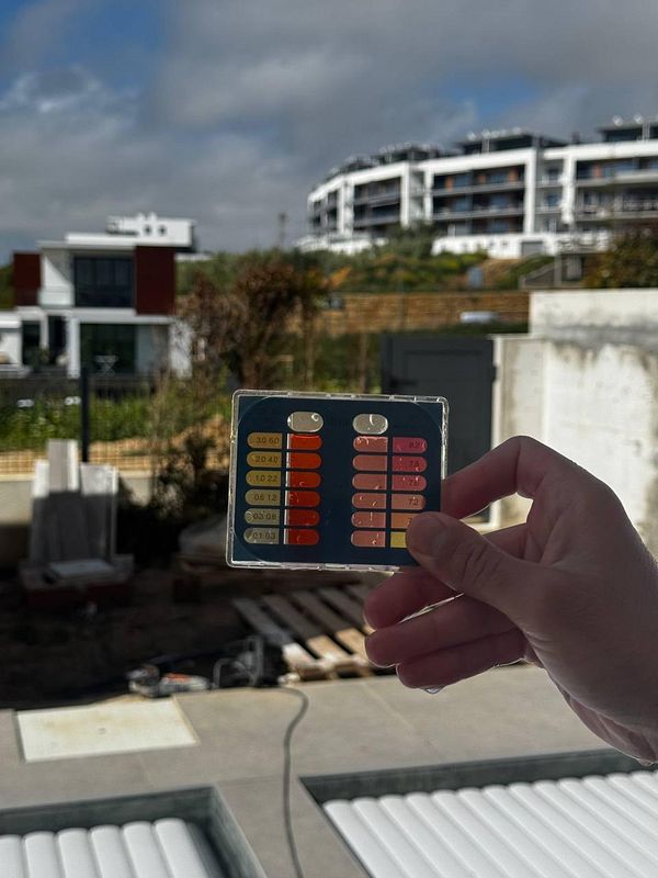 A hand holds a pH test strip in front of a jacuzzi area, with modern buildings in the background.