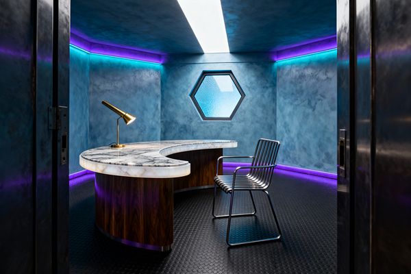A futuristic home office with smoky blue plaster walls and a hexagonal frosted window. A curved desk with a glowing backlit onyx top and walnut base sits on black coin-pattern rubber flooring, paired with a ribbed stainless-steel chair and a brass cone desk lamp. Cyan and purple LED cove lighting and a central linear ceiling light create a moody, space-age atmosphere.