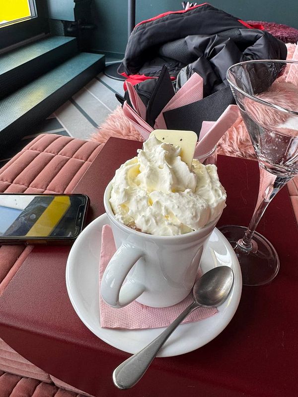 A cozy café scene featuring a cup of whipped cream topped drink and a glass, with a phone and ski gear in the background.