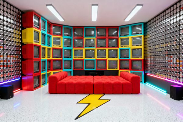 A media room features a floor-to-ceiling corner wall of vintage CRT televisions set into bold color-block frames in red, yellow, and turquoise. A low, plush red modular sofa sits on a white speckled floor with a yellow lightning-bolt inlay. The side walls are covered with rows of metallic can lids and stacked soup cans, while cyan and magenta LED strips wash the baseboards beneath linear ceiling lights, with black speakers and AV cabinetry grounding the setup.