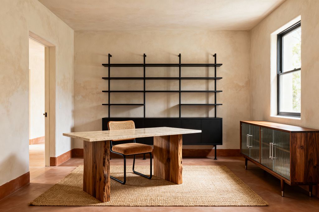 A serene home office with a light stone-top desk on live-edge wood legs centered on a jute rug. Behind the desk, a matte-black wall-mounted shelving system with a low credenza spans the wall. A black-framed window brightens warm limewashed beige walls and terracotta-toned floors. A caned-back chair with a tan upholstered seat sits at the desk, while a walnut sideboard with fluted glass doors and metal pulls lines the right wall; a doorway opens on the left.