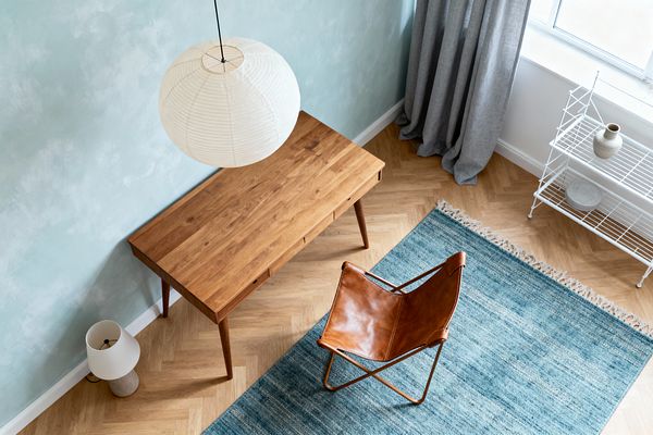 Top-down view of a Scandinavian home office with a slim oak writing desk against a pale blue textured wall. A cognac leather sling chair sits on a muted blue flatweave rug over light oak herringbone flooring. On the right, a white wire shelving unit holds a ceramic vase and plate beside a window dressed with gray linen curtains. A round white rice paper pendant hangs above, and a small table lamp with a stone base and fabric shade sits on the floor.