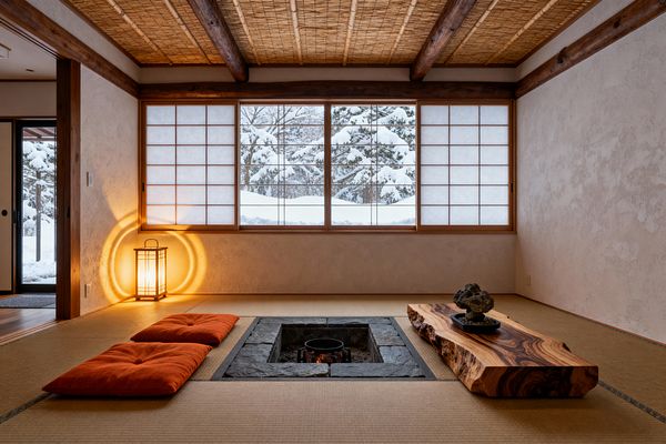 A minimalist Japanese living room with tatami flooring centers on a recessed irori hearth lined in dark stone and holding a small cast-iron pot. To one side lies a low live-edge wood table with a stone display; opposite sit two burnt-orange zabuton cushions. Shoji screens fill the rear wall, revealing a snowy pine landscape, while a wood-framed andon lantern glows beside textured plaster walls. The ceiling is woven bamboo with exposed timber beams.