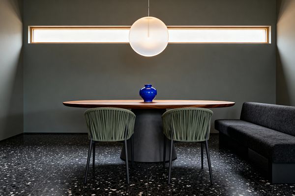 A modern dining room features an oval walnut table on a gray pedestal, flanked by two sage green rope-back chairs and a charcoal upholstered bench. A large frosted globe pendant hangs centered before a narrow horizontal clerestory window. The room has matte gray-green walls and black terrazzo flooring with white flecks; a bright cobalt vase sits on the tabletop.