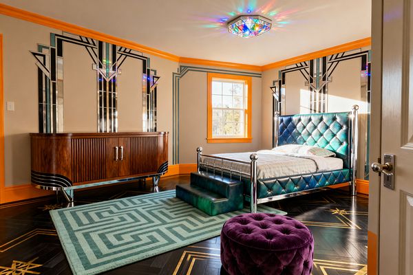 A kids’ bedroom in Art Deco style featuring a teal diamond‑tufted bed with chrome posts and an upholstered step before mirrored geometric wall panels. The space includes black herringbone wood flooring with gold inlays, a mint maze-patterned area rug, a purple tufted ottoman, and a walnut credenza on chrome legs. Beige walls with teal linework are framed by bright orange trim, and a jewel-like ceiling fixture with colored glass casts rainbow reflections.