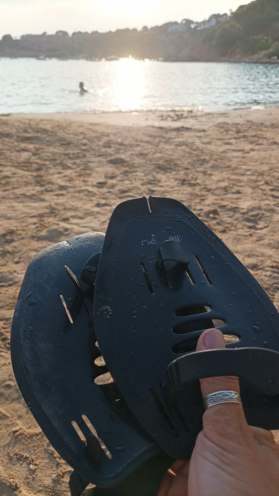 A person holds a pair of swim fins while sitting on a sandy beach with a view of the water and a swimmer in the background.