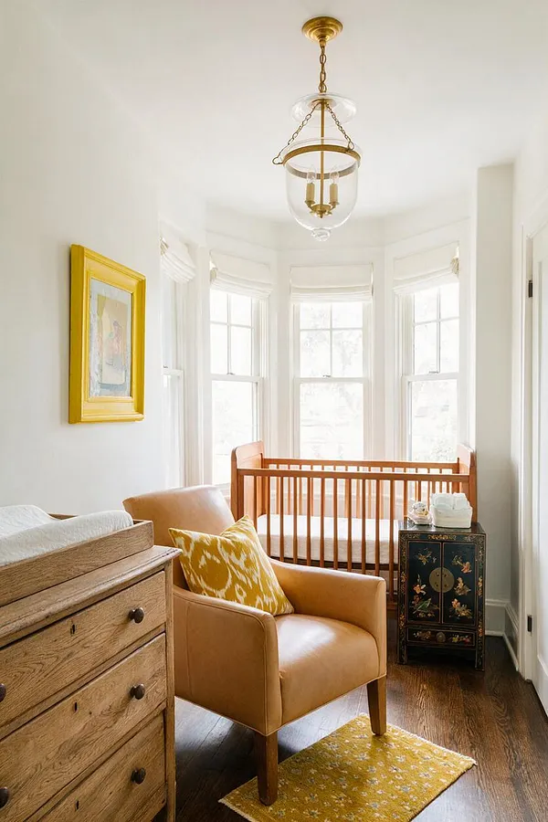 A bright eclectic nursery with a wood crib centered under bay windows, a tan lounge chair with a yellow patterned pillow, and a carved black side cabinet beside it. A natural wood dresser with a changing pad sits in the foreground, while a brass-and-glass pendant light and a gold-framed picture add warm vintage character.