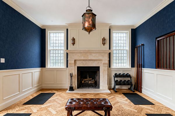 A refined home gym with a limestone fireplace centered between two leaded-glass windows, accented by brass sconces and a patinated lantern pendant. Deep navy wallcovering sits above cream paneled wainscoting; herringbone cork flooring holds black exercise mats and a compact dumbbell rack. A tufted brown leather bench anchors the foreground, creating a classic, light-filled workout space with traditional millwork and balanced symmetry.