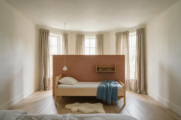 A bright Scandinavian-style sunroom with white walls, wide plank wood floors, and tall windows dressed in soft beige curtains. In the center sits a simple light wood bed with white bedding, a terracotta divider behind it, a small wall shelf, and a blue throw blanket adding a muted accent.