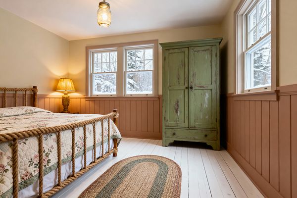 A cozy farmhouse kids' bedroom features a rope-framed bed with a floral quilt and a brass table lamp on a small nightstand. Clay-painted vertical wainscoting lines the lower walls beneath creamy paint, a distressed sage green armoire anchors the corner, and white painted plank floors hold an oval braided jute rug; two double-hung windows reveal snowy trees while a glass mason-jar pendant casts warm overhead light.
