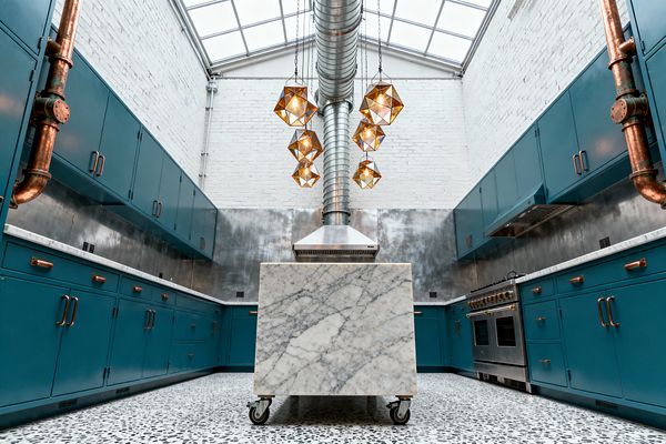 A skylit industrial kitchen features white-painted brick walls, teal flat-panel cabinets with copper pulls, and stainless-steel backsplash panels. An exposed galvanized duct drops to a wall-mounted stainless hood, flanked by geometric amber-glass pendants. A cube-shaped marble island on casters sits on a black-and-white terrazzo floor, with a pro-style stainless range at right and exposed copper plumbing risers on both side walls.