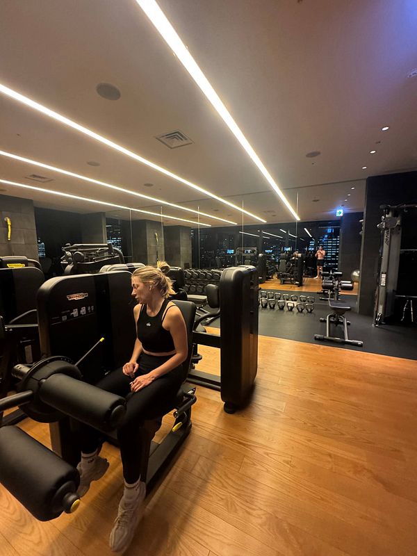A woman is seated on a gym machine in a modern fitness center with a view of the city skyline.