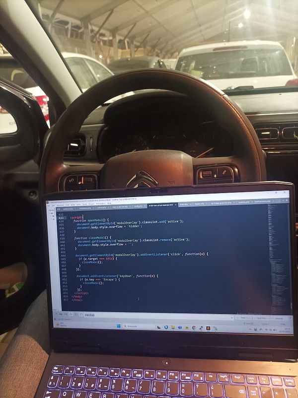 A laptop displaying code is positioned on the lap of a person sitting in a car, with a view of a ferry terminal in the background.