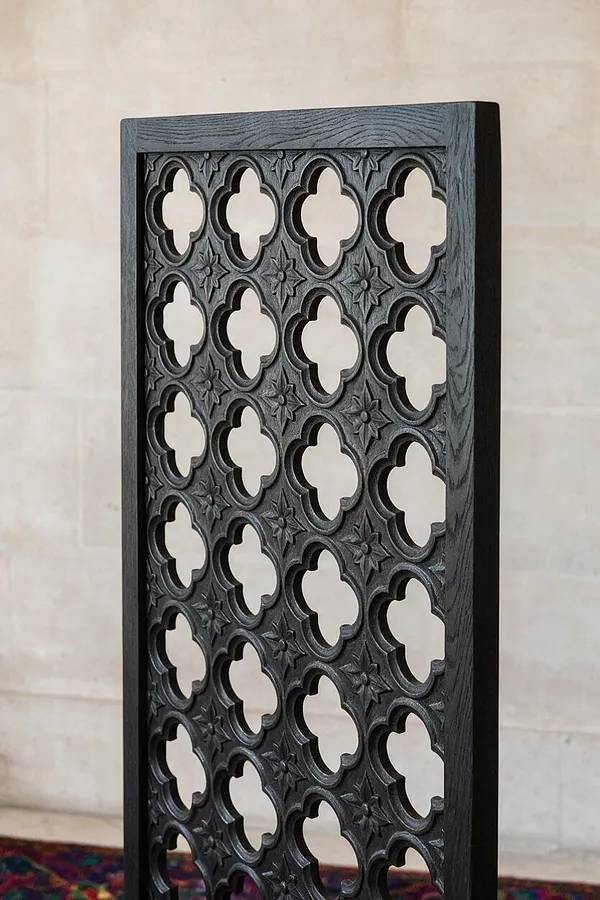 A tall rectangular decorative panel in dark black wood, featuring repeated Gothic quatrefoil cutouts and carved star motifs. The openwork design stands against a pale wall, with a glimpse of patterned carpet at the base.