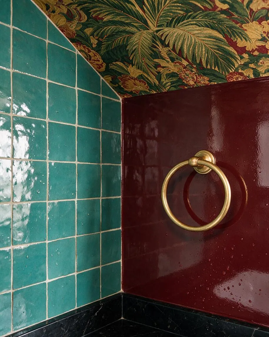 A close-up of a maximalist bathroom corner with glossy teal square wall tiles, a deep burgundy lacquered surface, and a brass towel ring. The ceiling is covered in dense tropical floral wallpaper, and a black stone ledge anchors the bottom of the composition.