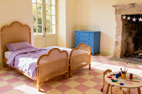 A sunlit children’s room with two scalloped rattan twin beds dressed in lavender linen on a pink-and-cream checkerboard tile floor. A rustic stone fireplace with a white star garland, a bright blue dresser, and a small round art table with paints sit beside tall French doors and pale plaster walls, creating a cozy, playful feel.