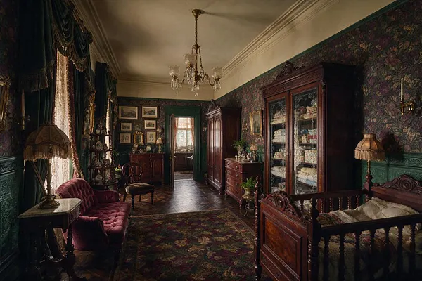 A richly decorated Victorian nursery with dark floral wallpaper, carved wooden cribs and cabinets, a patterned rug, and heavy drapery framing tall windows. Warm brass lighting, antique furnishings, and deep green trim create a moody, elegant atmosphere.