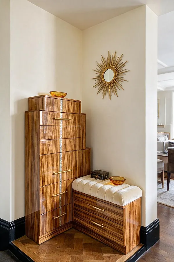 A compact Art Deco entryway features a tall striped wood cabinet with brass handles beside a matching cushioned bench in cream upholstery. A gold sunburst mirror hangs above, with amber glass bowls adding warmth against soft beige walls and dark baseboards.