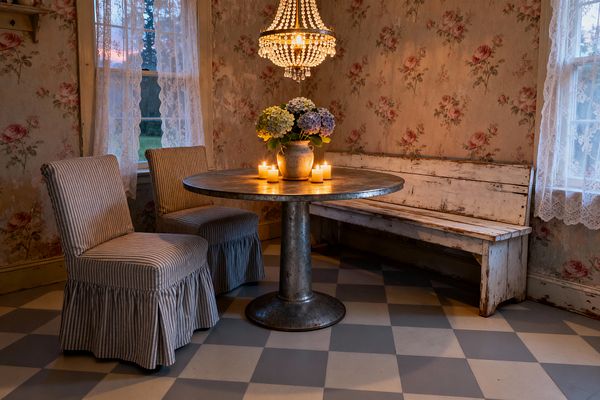 A cozy dining nook features a round galvanized metal pedestal table surrounded by two blue-and-ivory ticking slipcovered chairs and a long distressed white bench. Faded rose-print wallpaper, lace-curtained windows, a crystal beaded chandelier, a gray-and-cream checkerboard floor, lit pillar candles, and a vase of pastel hydrangeas create a softly aged, romantic Shabby Chic atmosphere.
