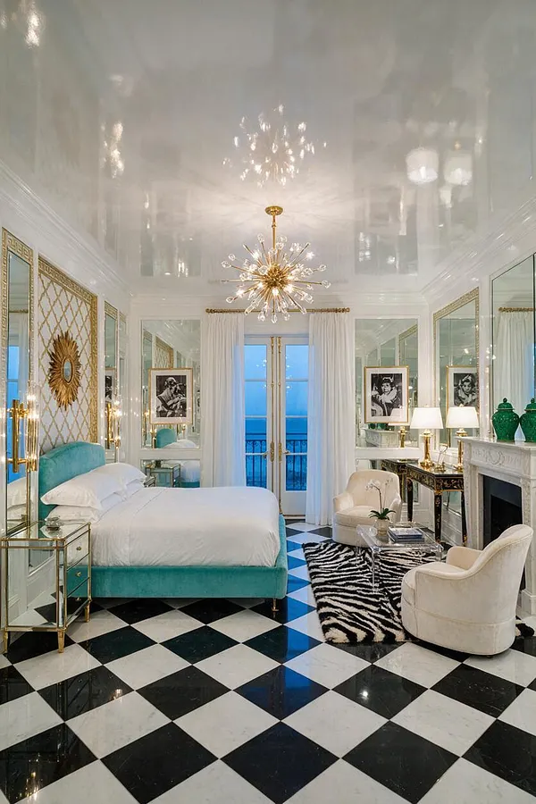 A luxurious master bedroom features a turquoise velvet bed, mirrored wall panels with gold lattice detailing, and a black-and-white checkered floor. A starburst chandelier, zebra rug, cream lounge chairs, and a white fireplace create a dramatic Hollywood Regency look.