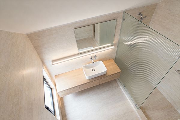 A minimalist bathroom clad in warm beige stone-look tiles features a floating light-wood vanity with a white rectangular vessel sink and wall-mounted chrome faucet below a wide frameless mirror. A slim linear LED runs along the wall, and a ribbed glass shower screen defines the walk-in area with a simple chrome shower set. A small black-framed window brings daylight, and integrated drawers keep the space uncluttered.