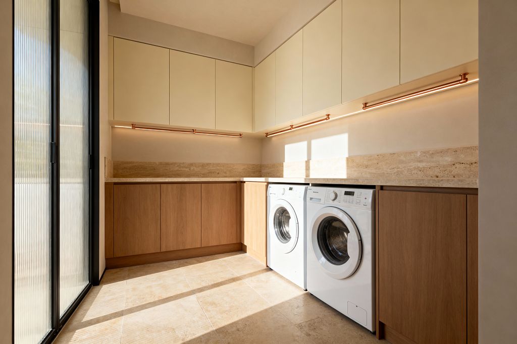 A bright contemporary laundry room features cream flat-panel upper cabinets and natural oak base cabinets surrounding a white front-loading washer and dryer. A continuous travertine countertop and backsplash meet large-format stone floor tiles, while warm LED light bars on copper-finish rails illuminate the workspace. A black-framed sliding door with fluted glass brings daylight, highlighting handleless cabinetry and a palette of beige, cream, oak, and white.
