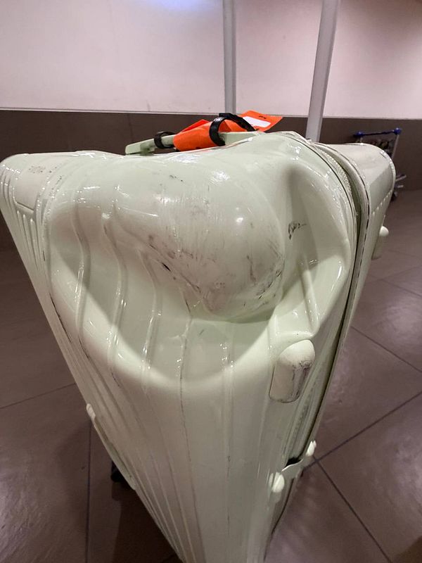 A heavily damaged black Rimowa suitcase placed on airport luggage belt with visible scratches, dents, and a broken handle.