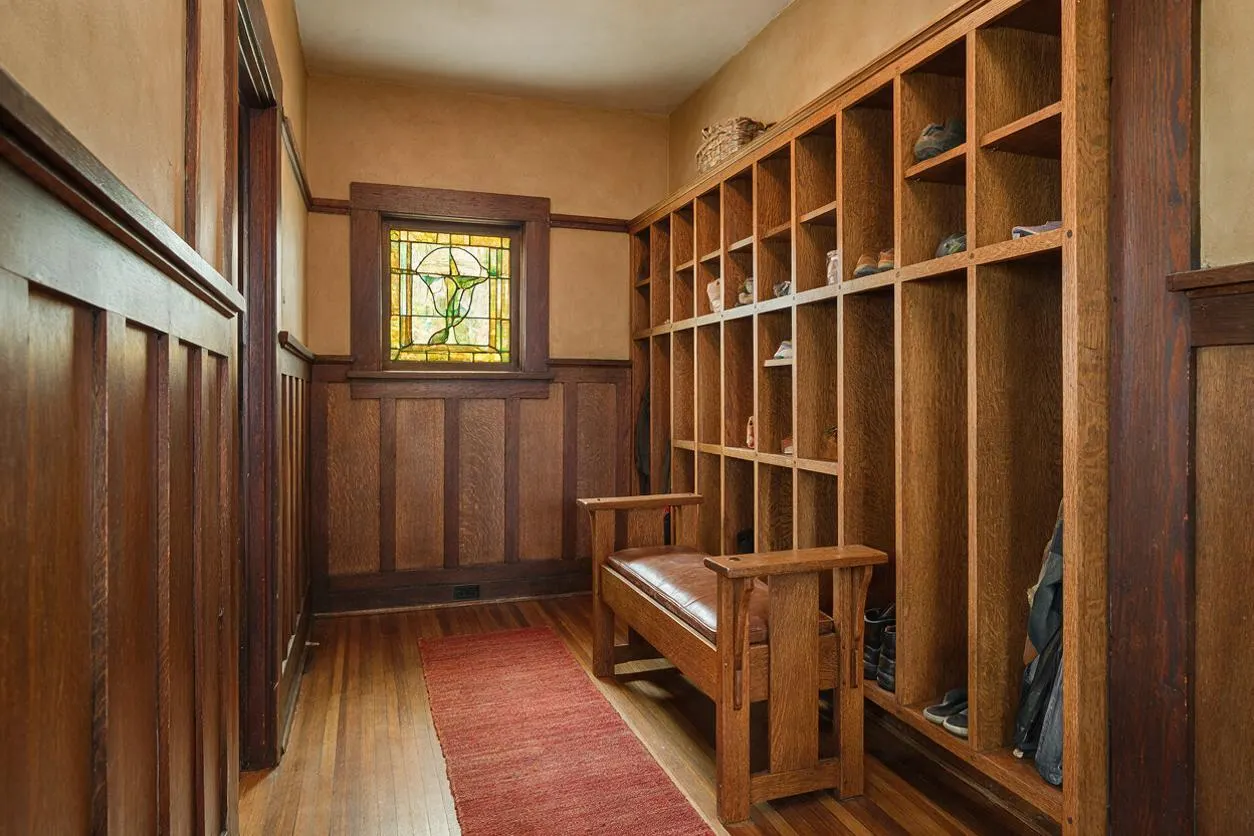 A narrow Craftsman-style mudroom with rich wood wainscoting, a stained glass window, and a long wall of open cubbies for shoes and storage. A wooden bench with a leather seat sits beside a red runner rug on the hardwood floor, creating a warm, organized entry space.