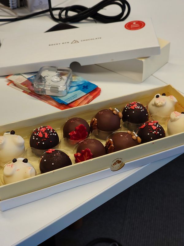 A box of pralines is displayed on a desk, accompanied by a chocolate box and some paperwork.