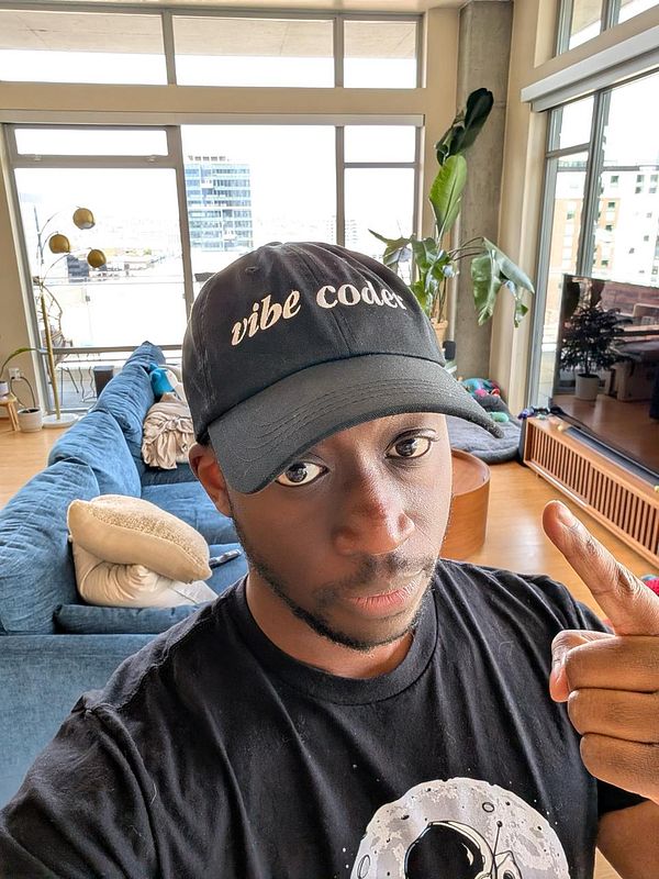 A person wearing a black 'vibe coder' hat poses in a bright, modern living space.