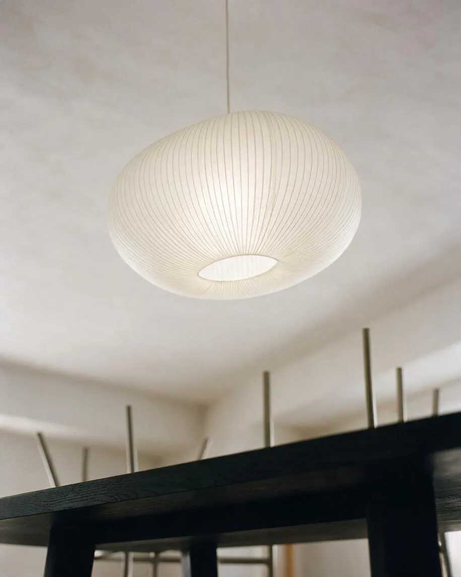 A modern interior ceiling light with a large oval paper lantern shade hanging from a thin cord. The soft ivory pendant has fine vertical ribbing and a circular opening on the bottom, set against a pale ceiling with a dark table edge visible below.