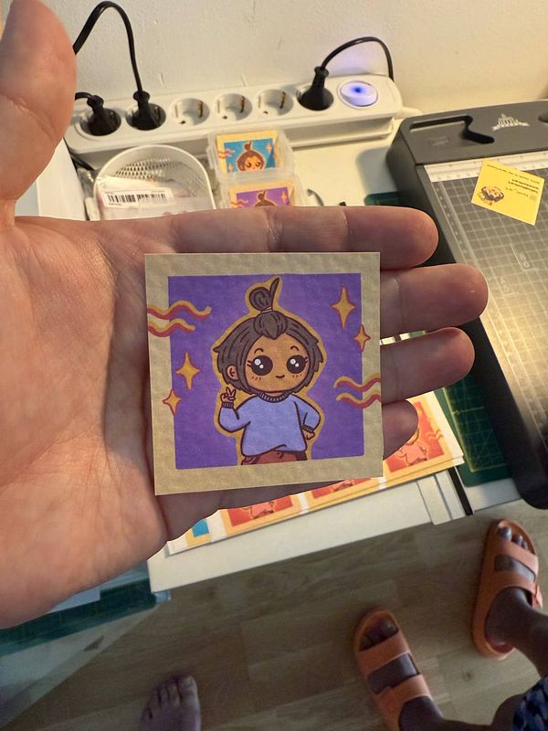 A hand holds a colorful illustration of a character with a playful pose.