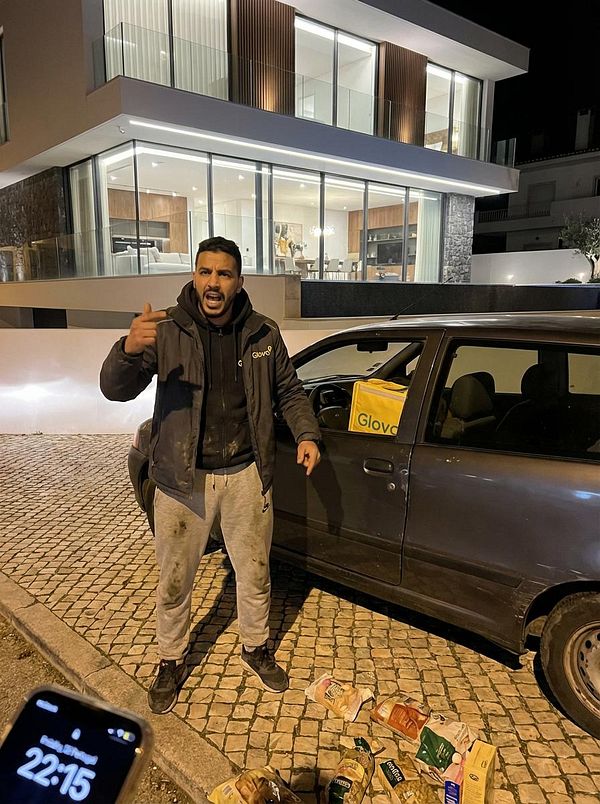 A man in a car beside a gate and a white wall in an outdoor residential area is shouting aggressively and throwing groceries.