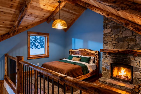 A rustic loft bedroom features slate blue walls, a knotty pine cathedral ceiling with exposed log beams, and a live-edge hardwood bed dressed in rust and forest green bedding. A fieldstone fireplace with a thick timber mantel burns brightly beside the bed, while a hammered brass pendant, honey-toned wood floors, log-and-iron railing, and a small window framing snowy pines create a warm, cabin-like atmosphere.
