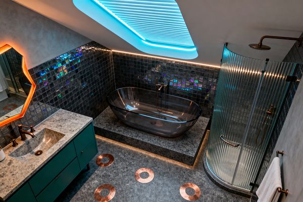 A futuristic bathroom under a sloped ceiling features a transparent smoked-glass tub on a gray stone platform, framed by iridescent dark mosaic wall tiles and warm brass fixtures. At right is a curved shower with fluted glass, while a teal vanity with stone countertop and a faceted mirror with orange LED backlighting face the tub; copper ring inlays punctuate the charcoal floor, and cyan cove lighting with warm perimeter LEDs sets a moody glow.