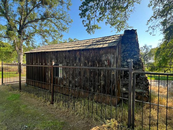 A rustic cabin surrounded by a black metal fence, set against a backdrop of trees and blue sky.