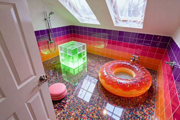 A whimsical attic bathroom with sloped ceiling and skylights features a glossy epoxy floor embedded with rainbow sprinkles and ombré ceramic wall tiles shifting from purple to pink to orange. A donut-shaped resin tub with faux sugar and sprinkles sits beside a glowing neon green acrylic cube and a pink macaron stool, while chrome wall taps and a handheld shower complete the playful, candy-themed space.