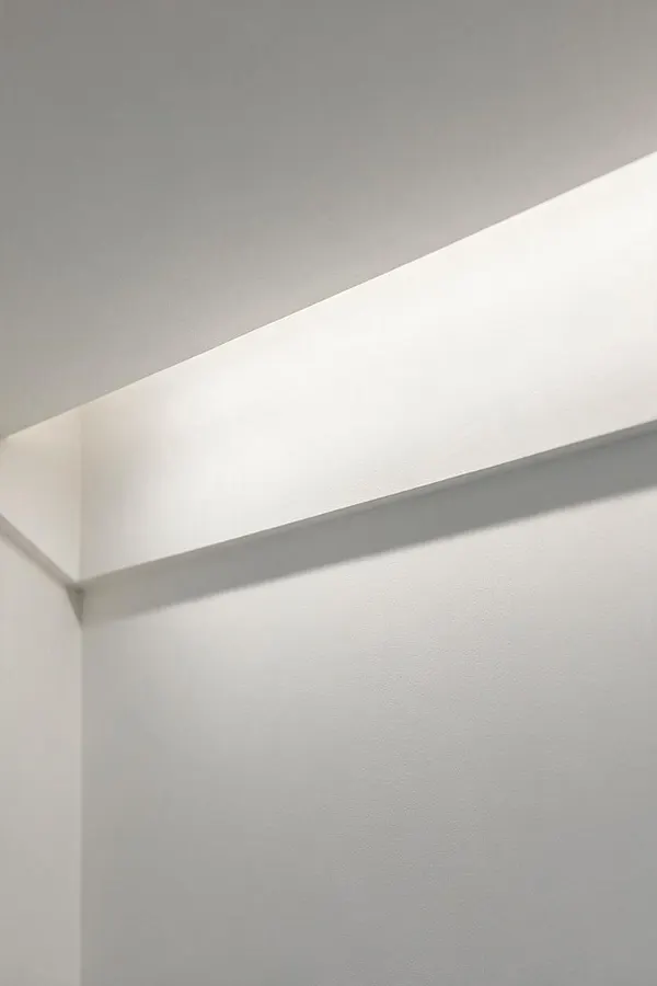 A close-up of a minimalist interior wall and ceiling corner painted in soft white and light gray. A clean horizontal architectural ledge casts a subtle shadow across the smooth surface, creating a simple geometric composition with no decoration.