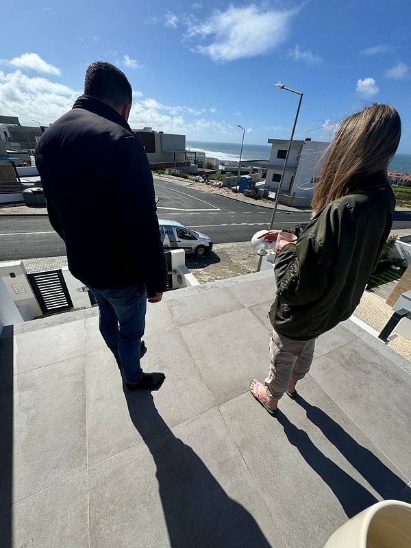 Two individuals stand on a balcony overlooking a coastal view.