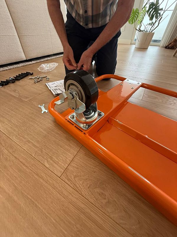 A person is assembling a heavy-duty carriage with a large wheel on a wooden floor.