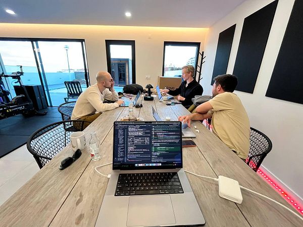 A cozy indoor coworking space with seven people working individually on laptops around a wooden table.