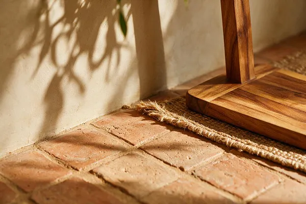 A close-up interior scene showing a terracotta tile floor with warm, earthy tones beside a wooden furniture base and woven jute rug edge. Soft sunlight casts leafy shadows on a textured wall, creating a calm bohemian atmosphere.