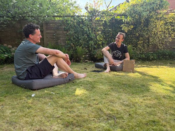 Two men are sitting on cushions in a garden, engaged in conversation.