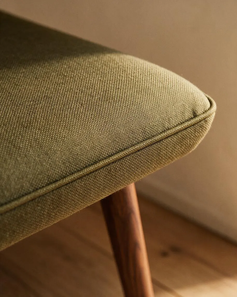 Close-up of a mid-century modern upholstered seat in olive green fabric with fine woven texture and tailored piping along the edge. A tapered walnut leg is visible beneath the cushion, with warm wood flooring and a neutral wall in the background.