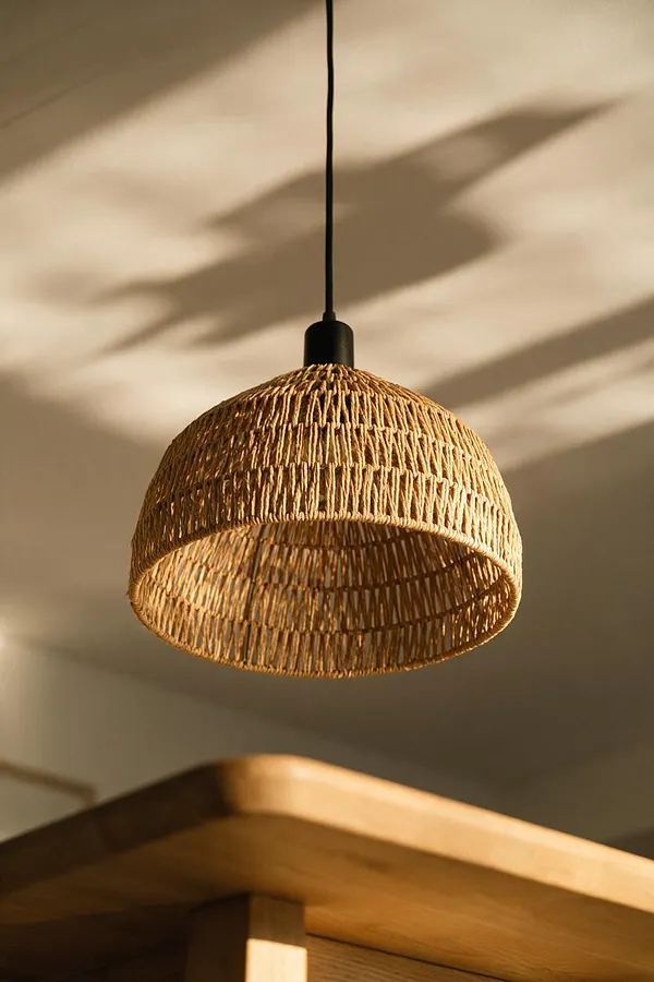 A close-up of a dome-shaped woven pendant light hanging from a black cord against a soft beige wall. Warm sunlight casts organic shadows across the ceiling, while a light wood furniture edge appears at the bottom of the frame.