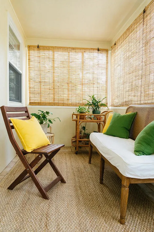 A small tropical front porch with bamboo roller shades on three sides, a woven jute rug, a wooden folding chair with a bright yellow cushion, and a cushioned bench with green and yellow pillows. Potted plants and a bamboo side table add a relaxed, airy feel.