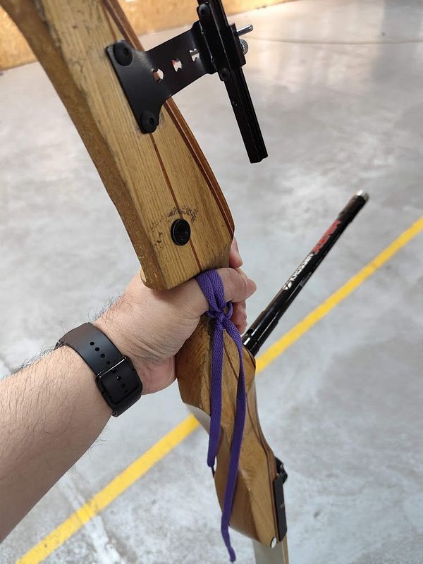 Person gripping a wooden recurve bow in an indoor archery range with a purple string loop tied around the wrist.