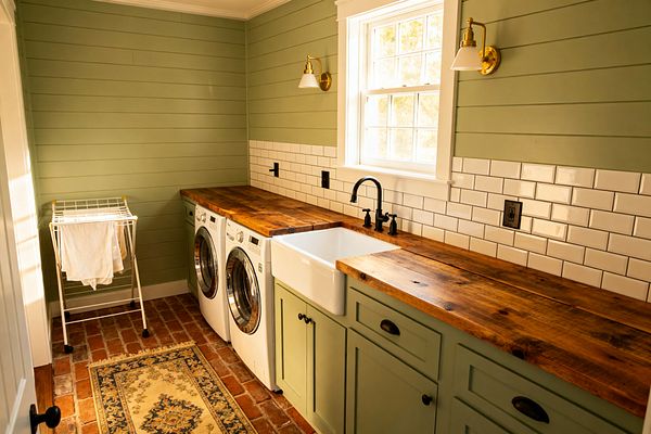 A farmhouse-style laundry room features sage green shiplap walls and matching cabinets topped with thick reclaimed wood countertops. A white apron-front sink with a matte black bridge faucet sits between a front-loading washer and dryer. Glossy white subway tile forms the backsplash beneath a window with white trim. The floor is red brick with a patterned rug, a folding drying rack stands at left, and brass sconces with white shades provide warm light.