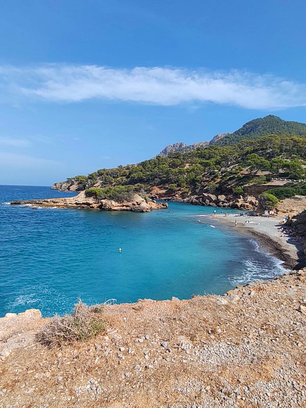 A scenic coastal cove with clear turquoise water, rocky shoreline, and a small pebbly beach surrounded by green hills under a clear blue sky.