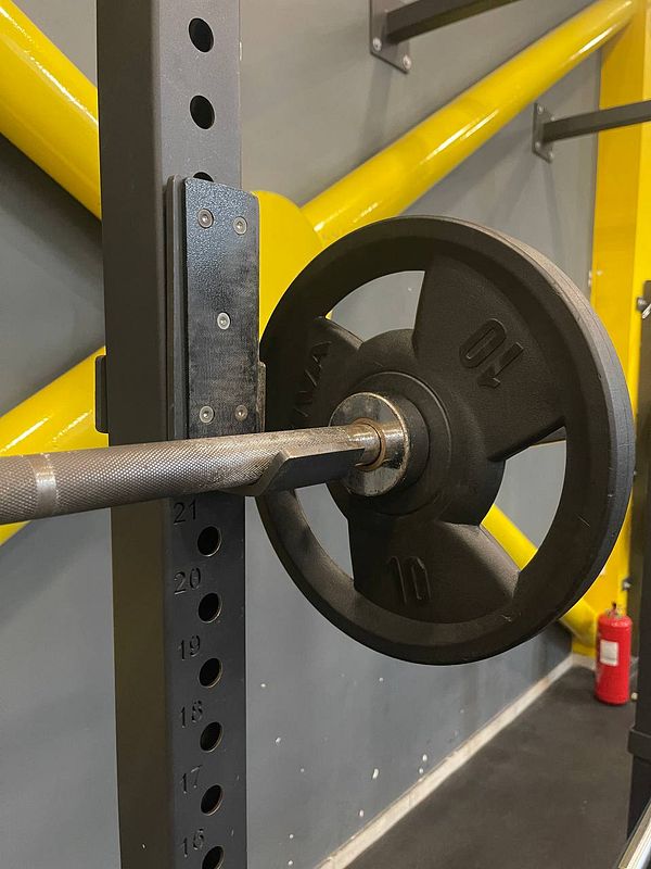 A close-up view of a weightlifting barbell with a 70 kg weight plate attached, positioned on a squat rack in a gym.